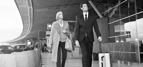 A Blacklane chauffeur takes a guest's luggage while guiding her to his vehicle at the arrivals gate of an airport. Both wear masks.