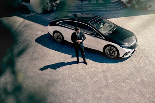 A suited Blacklane chauffeur stands outside a two-tone Mercedes EQE limo outside a villa.