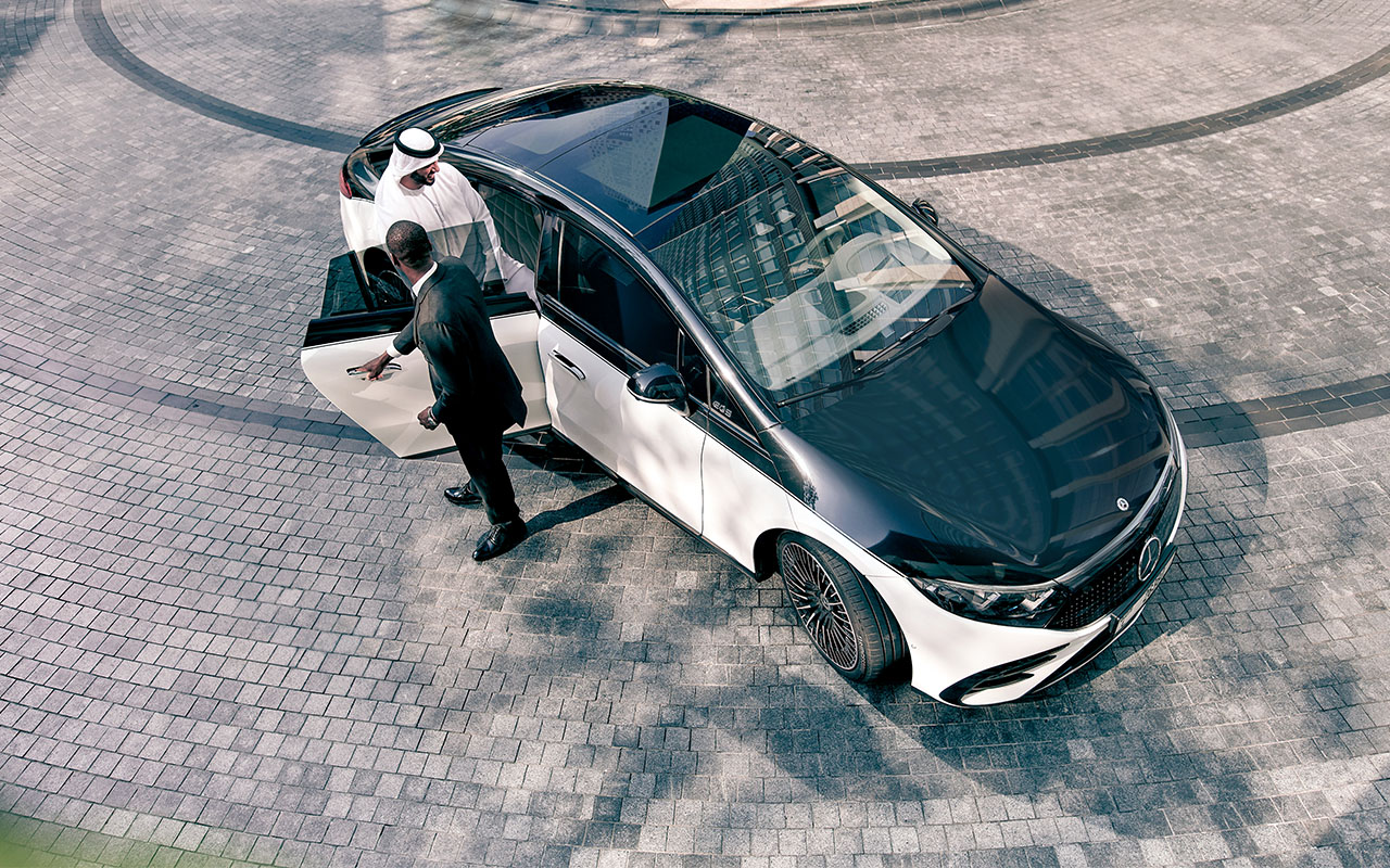 A chauffeur holds the door of his Mercedes-Benz EQS open for his guest.