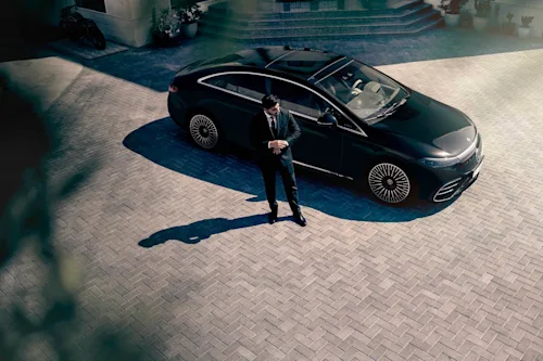 A Blacklane chauffeur adjusts the cuffs of his suit, standing in front of his Tesla.