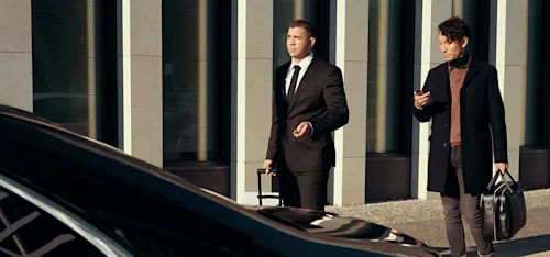 A Blacklane chauffeur guides an airport transfer customer to his waiting limo while carrying his luggage.