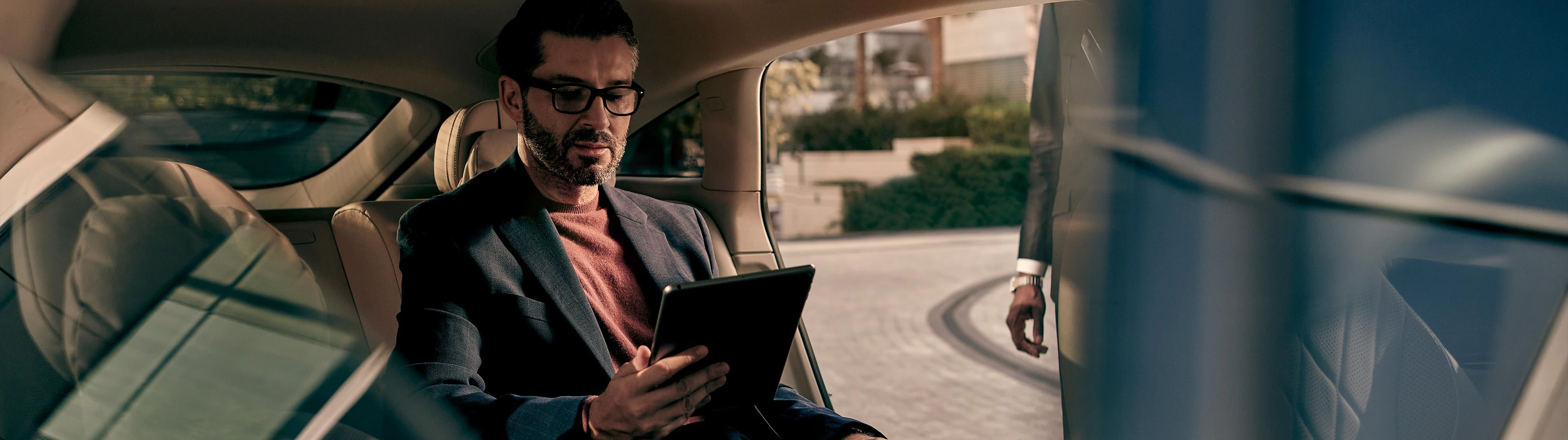 A man using a tablet sits in the back seat of a Mercedes-Benz while his chauffeur closes the door after him.