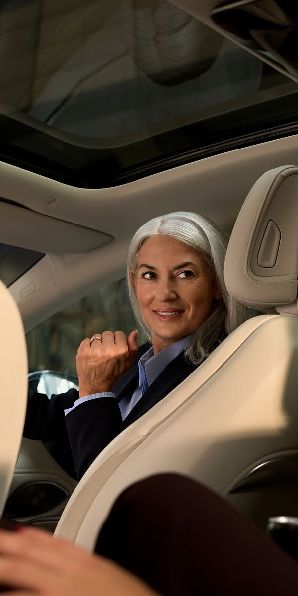 A Blacklane chauffeuse turns around smiling in the driver's seat to look at their passenger in the back seat of a limousine.