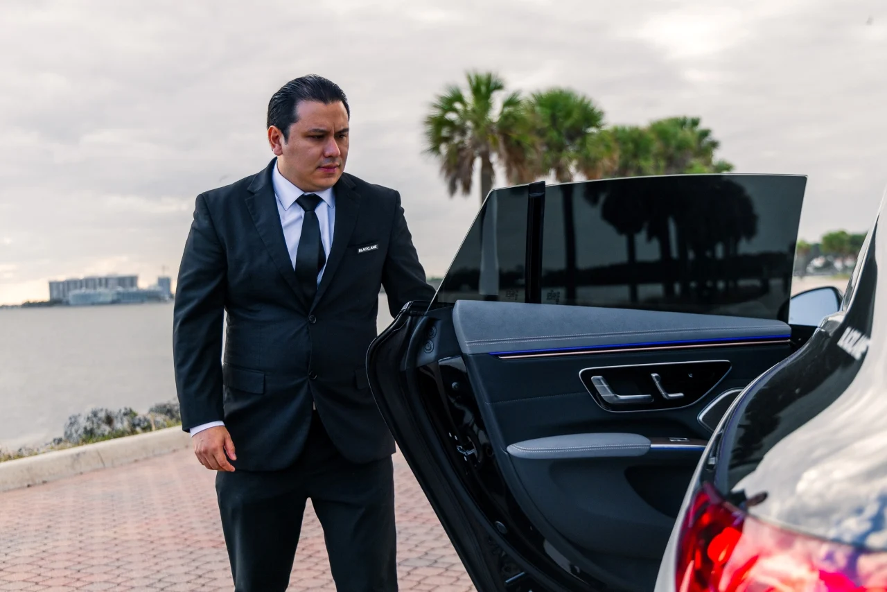 A Blacklane chauffeur in Miami opens a limo door with the sea and palm trees in the background.