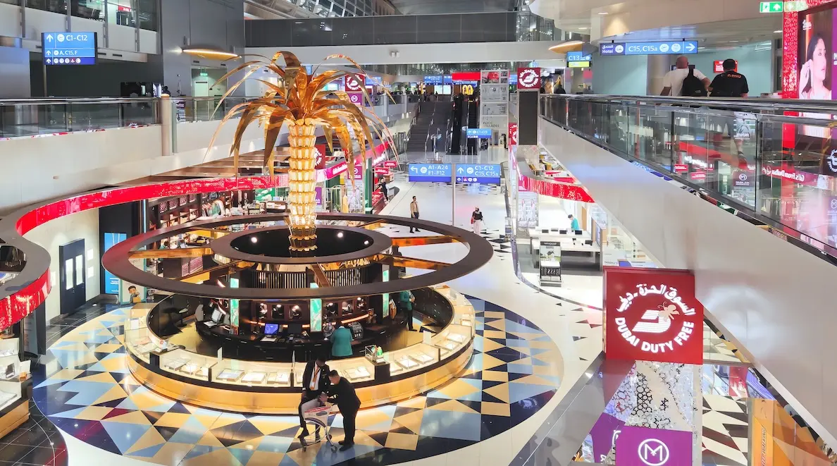 Modern interior of Dubai International Airport Terminal 3 showing shopping area with signature decorative artificial palm trees and rows of empty passenger seating.