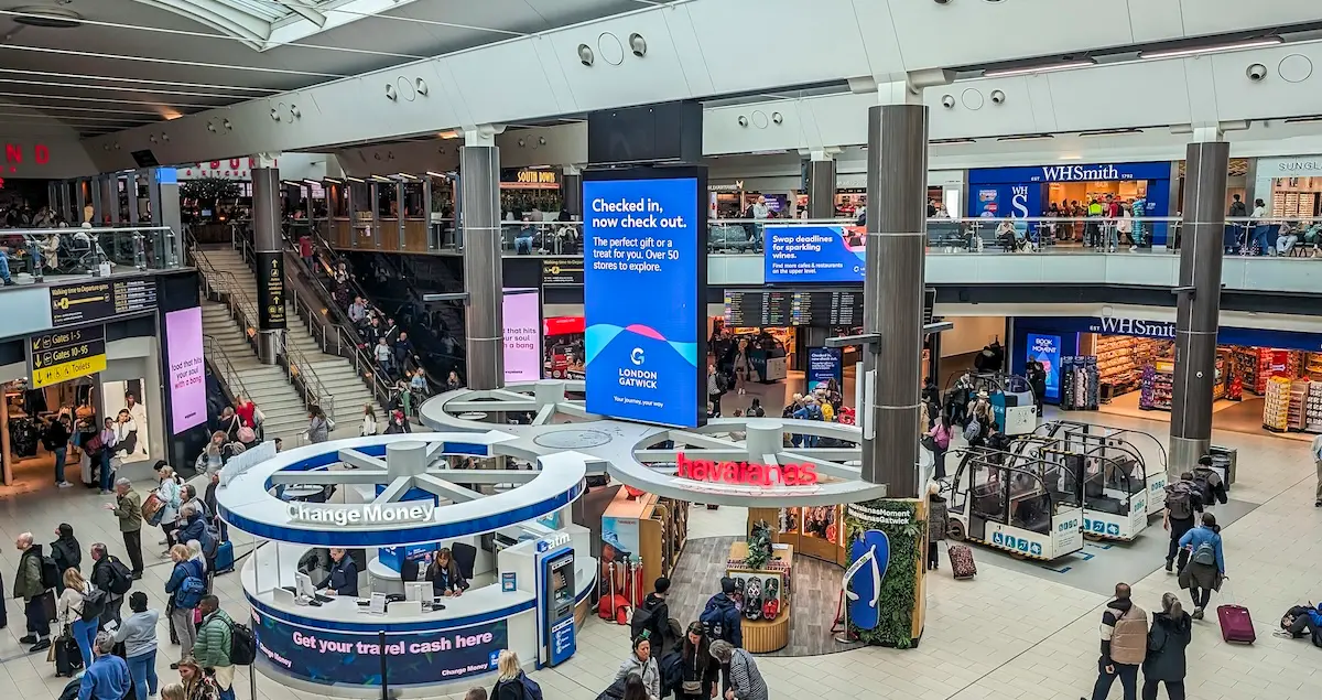 Interior overview of London Gatwick Airport North Terminal with shops, money exchanges, and passengers walking by.