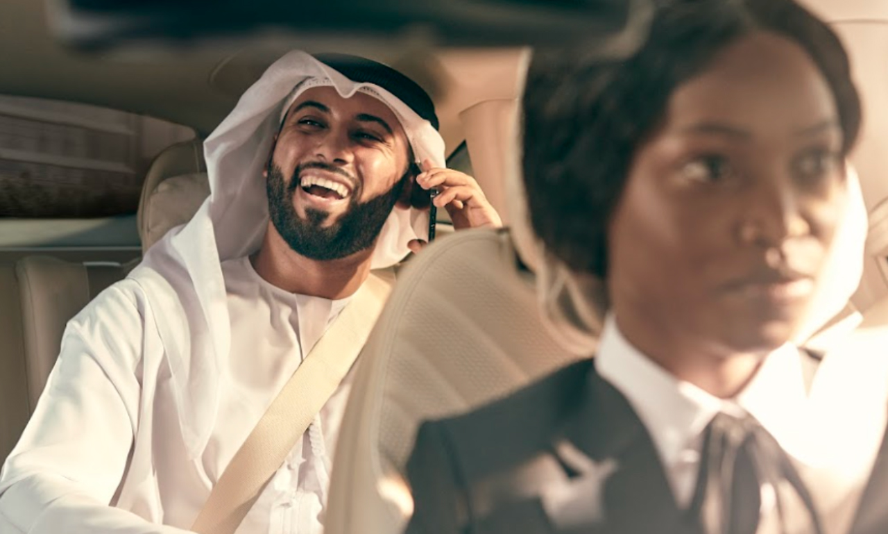 Female chauffeur focuses on driving while man talks on his phone in the backseat.