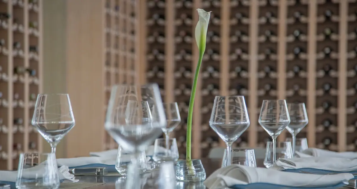 An elegant private dining table covered in glassware with a single lily centerpiece and racks of wine bottles on the walls.