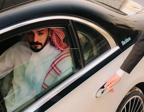 A man in traditional Saudi dress sits in the back seat of a Blacklane limo, as the chauffeur's suited hand reaches for the door.