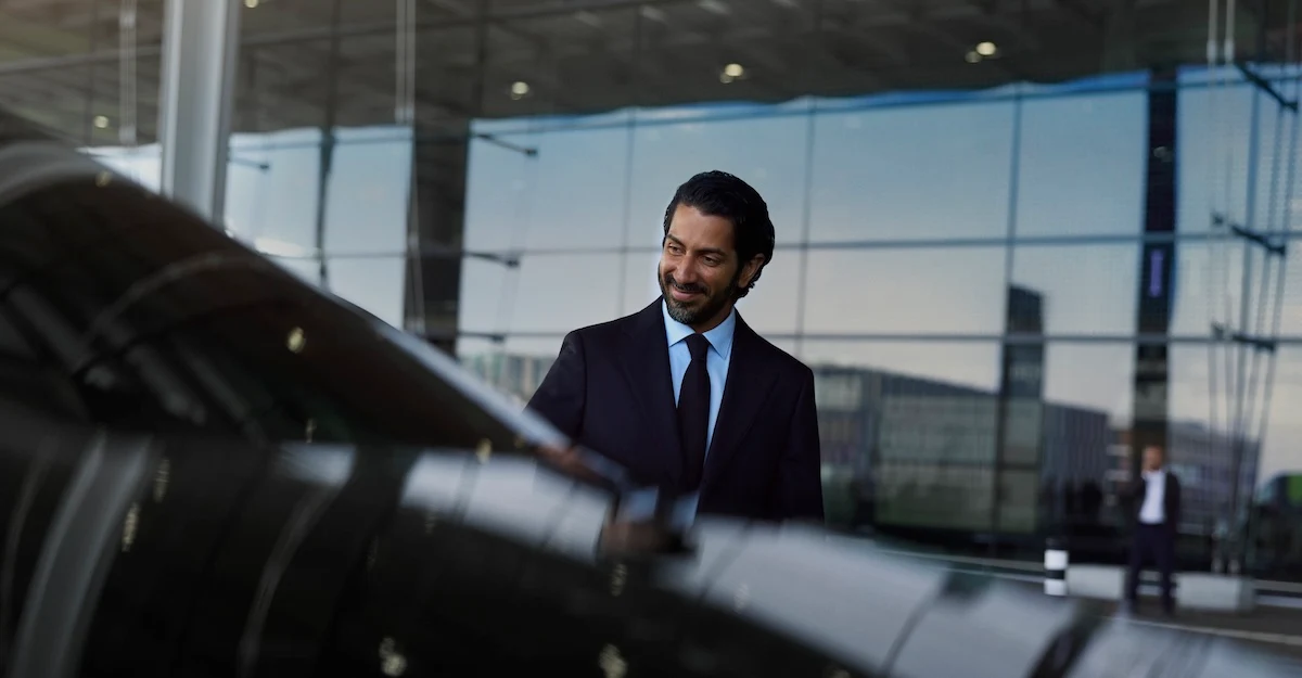 A chauffeur in a suit stands smiling next to a black vehicle.