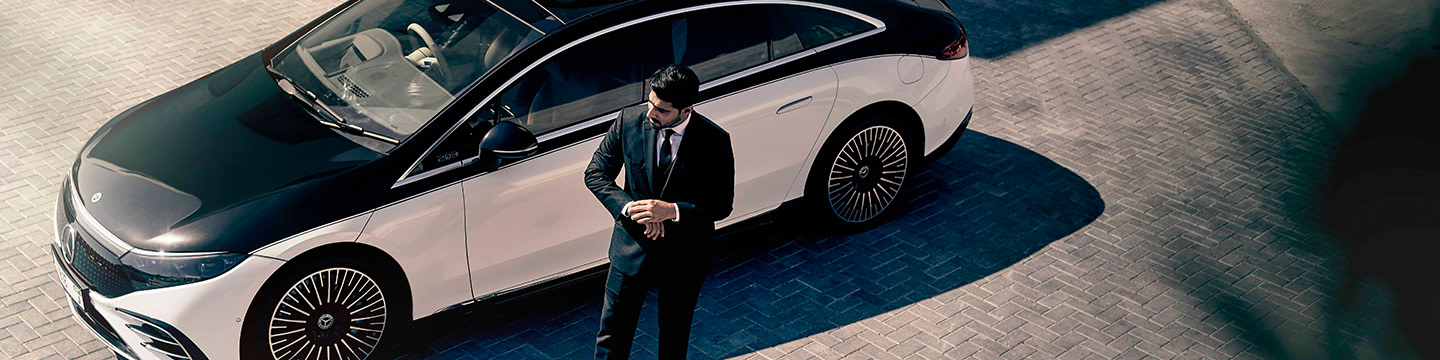 A Blacklane chauffeur in a suit stands outside of his two-tone Mercedes-Benz EQS.