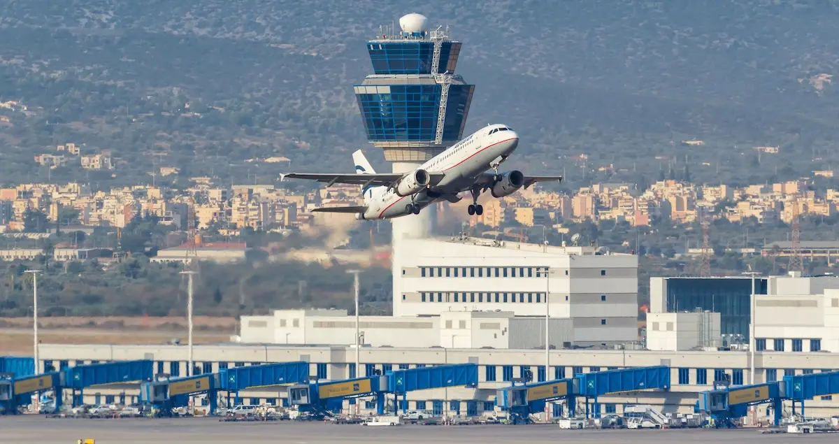 A plane takes off from Athens AIA Airport showing the terminal building, skywalks, and the control tower behind it.