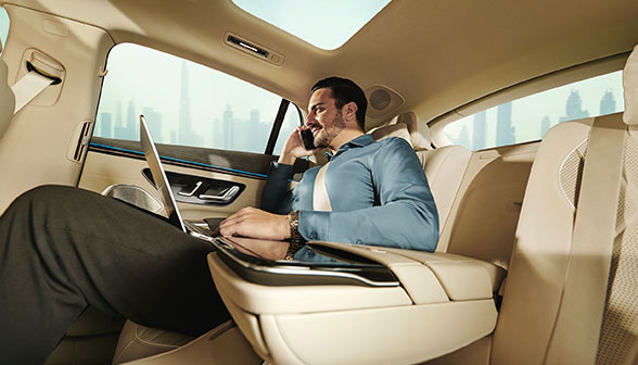 Business man sits in the backseat of a Mercedes EQS with a laptop rested on his lap while talking on a cellphone.