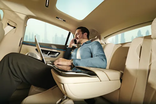 Ein Geschäftsmann sitzt auf dem Rücksitz eines Mercedes EQS mit einem Laptop auf dem Schoß und telefoniert mit einem Handy.