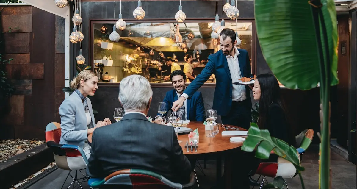 A waiter serves 4 businesspeople at a private dinner, with an open kitchen in the back.