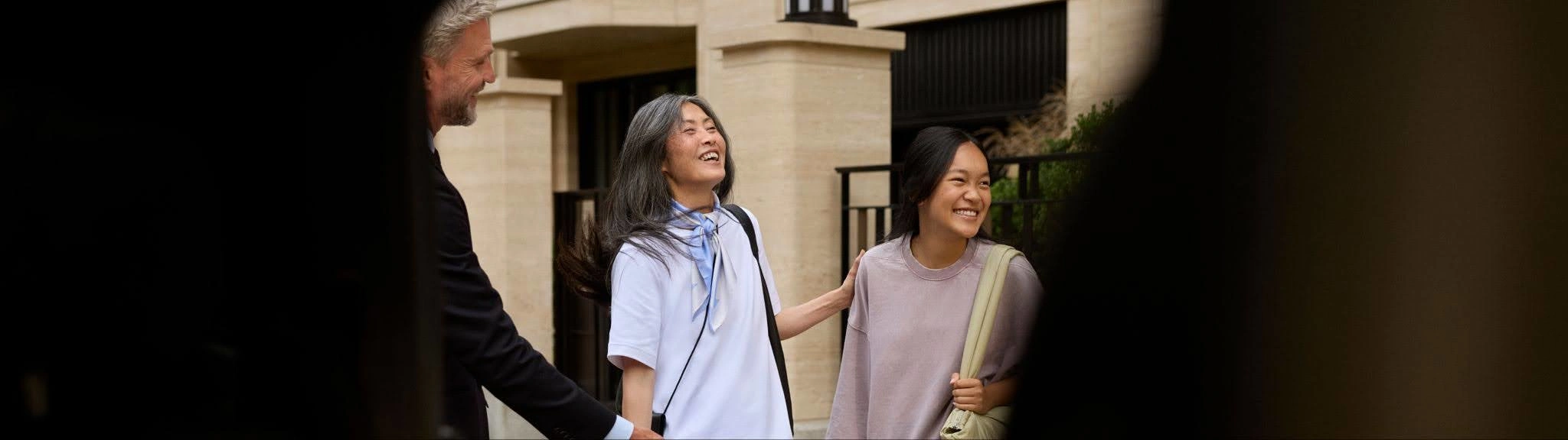 A woman and daughter stand laughing with luggage beside a suited Blacklane chauffeur who is taking their bags to the car.