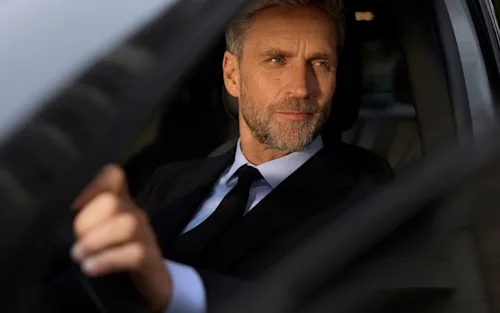 A Blacklane chauffeur with salt and pepper hair looks out the open window of his car while at the wheel.