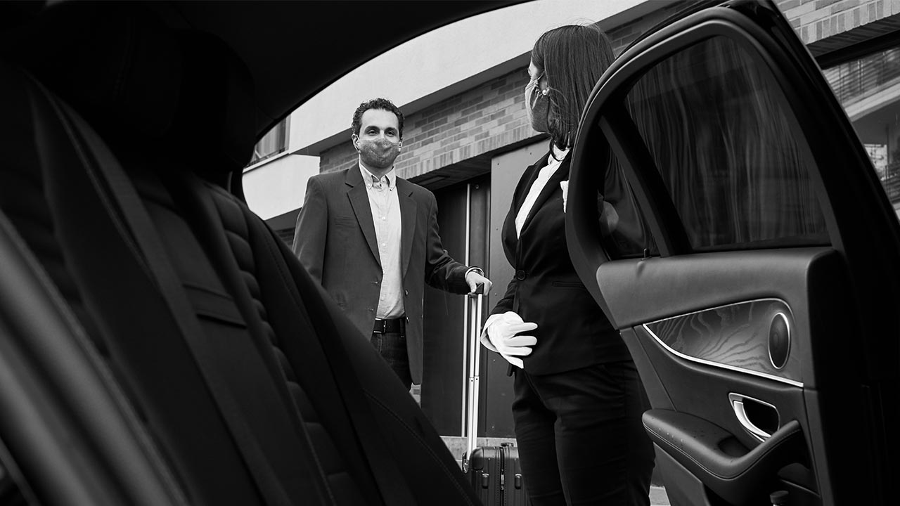 A real Blacklane chauffeur wearing gloves and a face mask opens the back door of a black car for a guest who is also wearing a mask and holding the handle of his luggage.