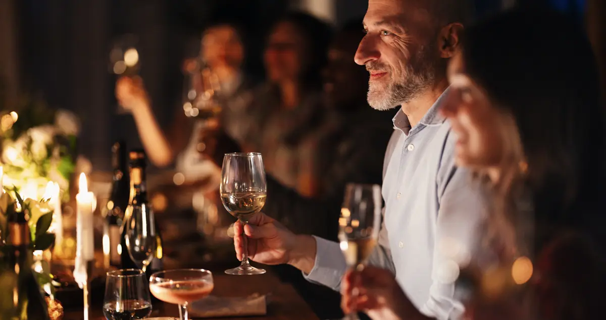 Some members of a private dining party holding their glasses up for a toast, with a candlelit table of food and drink in front of them.