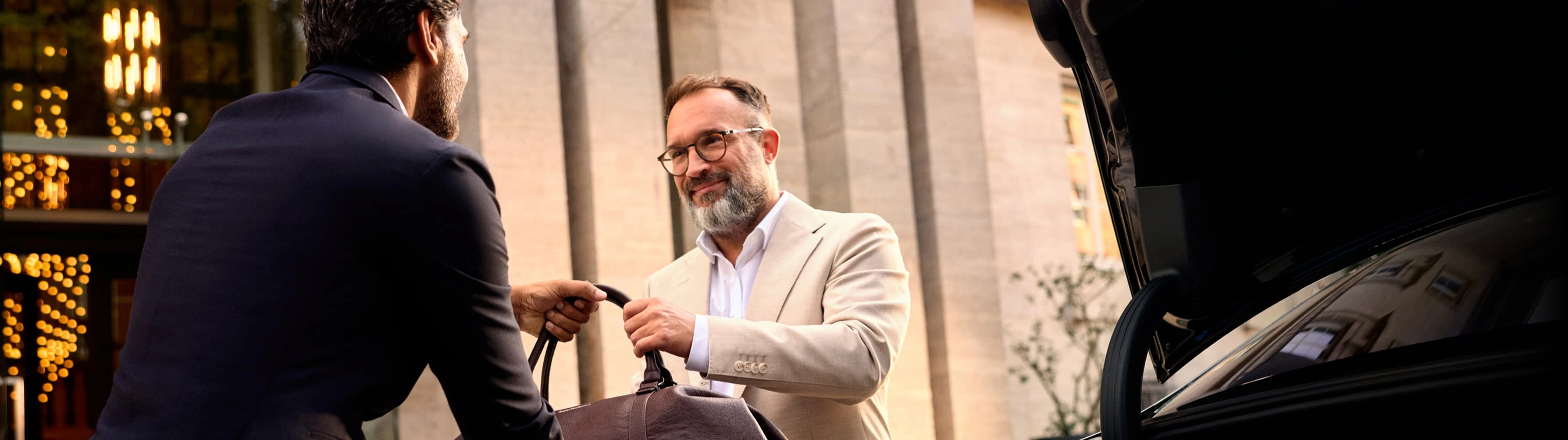 A well-dressed traveler takes his bag from a suitcase outside a luxury hotel, with a Blacklane limo in the left foreground.