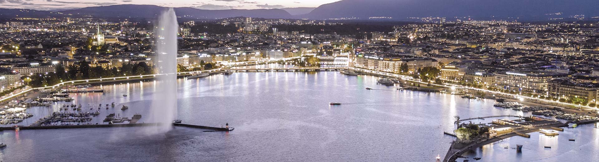 The beautiful cityscape of Geneva at night with an active fountain in the foreground.