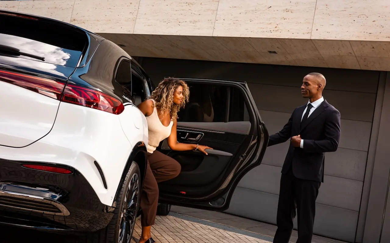 A woman steps out of a Blacklane two-tone Mercedes limo as a smartly dressed chauffeur holds open the door for her.