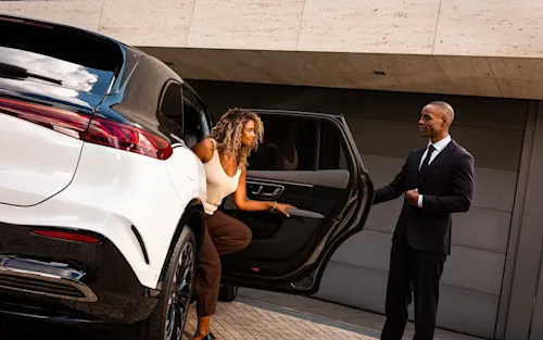 A woman steps out of a Blacklane two-tone Mercedes limo as a smartly dressed chauffeur holds open the door for her.