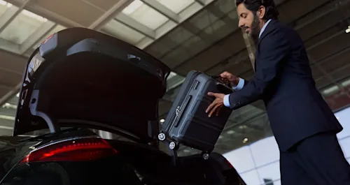 A Blacklane chauffeur places a luggage case in the trunk of a black Mercedes limousine outside an airport terminal.