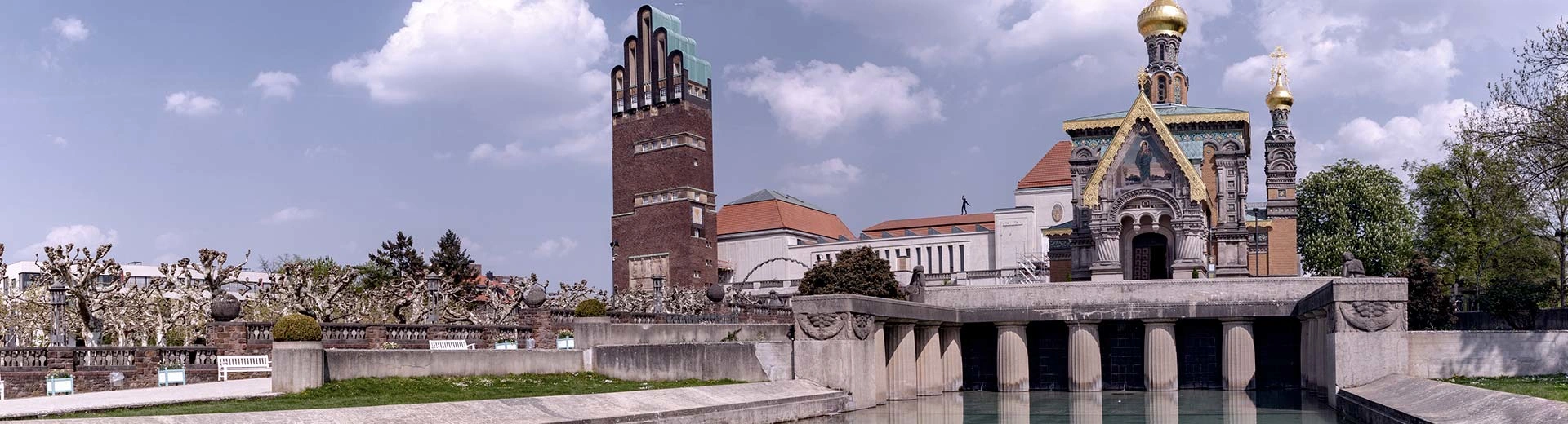 The historic churches and buildings of Darmstadt sit next to a canal on a clear and sunny day.