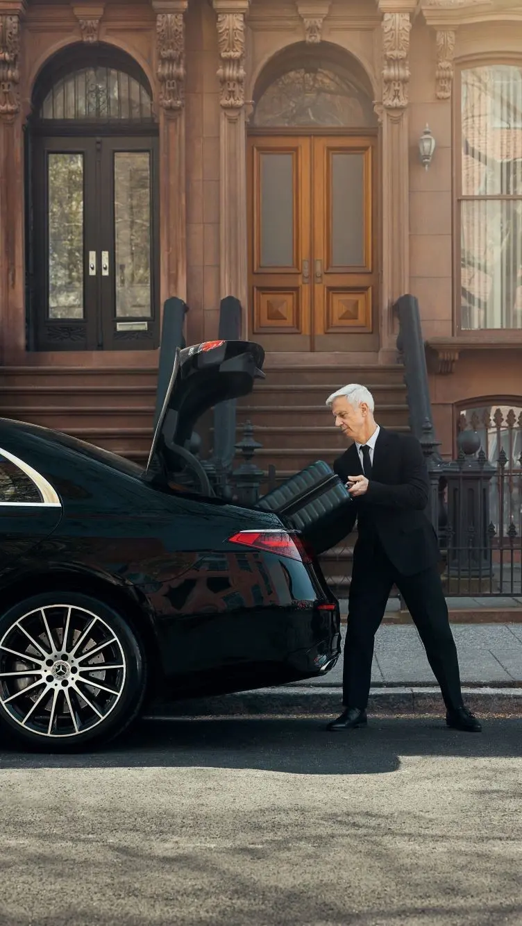 Un chauffeur charge des bagages à l'arrière d'un véhicule, devant deux maisons de ville emblématiques en pierre brune.