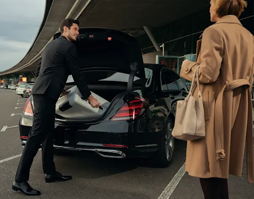 A Blacklane chauffeur packs a woman's bags into the back of the vehicle as she waits nearby.