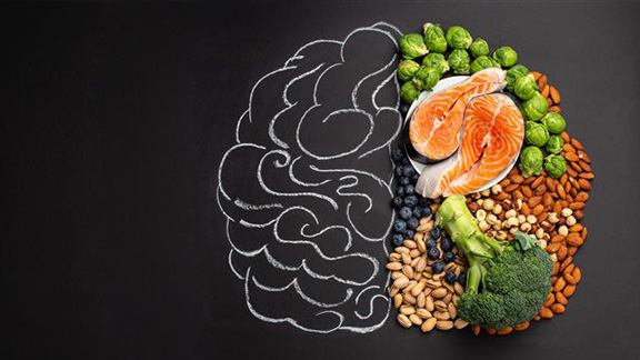 A chalk-drawn brain is half-filled with healthy foods: salmon, nuts, Brussels sprouts, broccoli, and blueberries, against a black background.