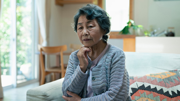 Elderly woman sitting on couch by window, looking off in a distance.