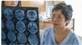 An older woman wearing glasses and a hospital gown looks at brain scan images, with a medical professional partially visible beside her.