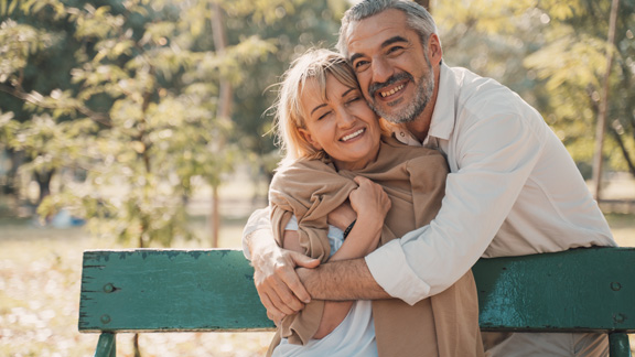 A happy couple hugging on a green bench smiling in the woods.