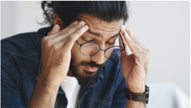 A man, wearing glasses, in a denim button up and white shirt. Holding his temples in pain with eyes closed.