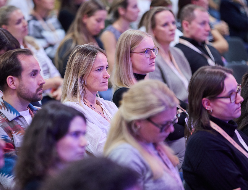 A diverse audience attentively listening at a conference, with attendees wearing lanyards seated in rows.