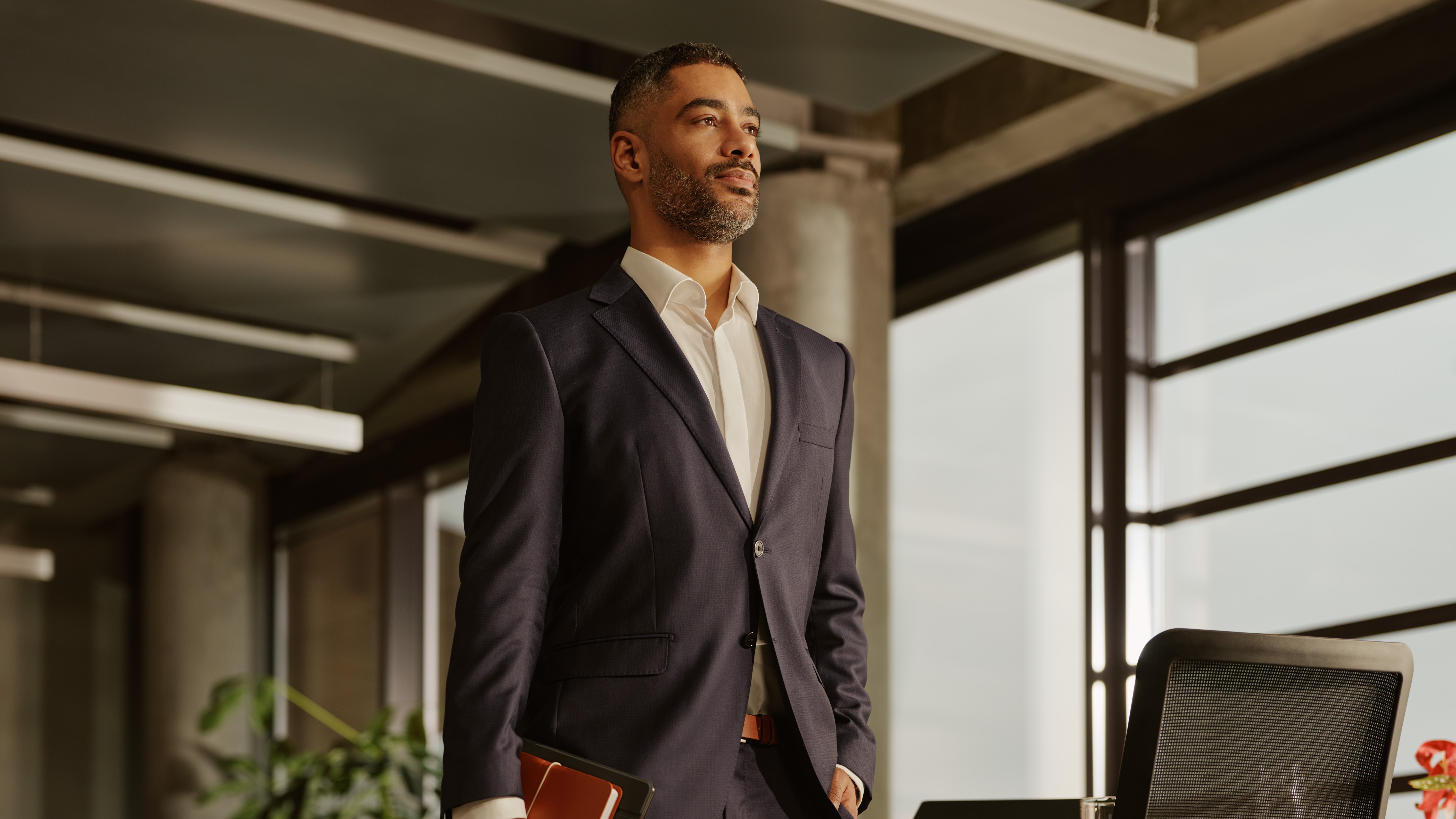 A confident businessman in a navy suit holds a tablet and notebook, standing thoughtfully in a modern office.