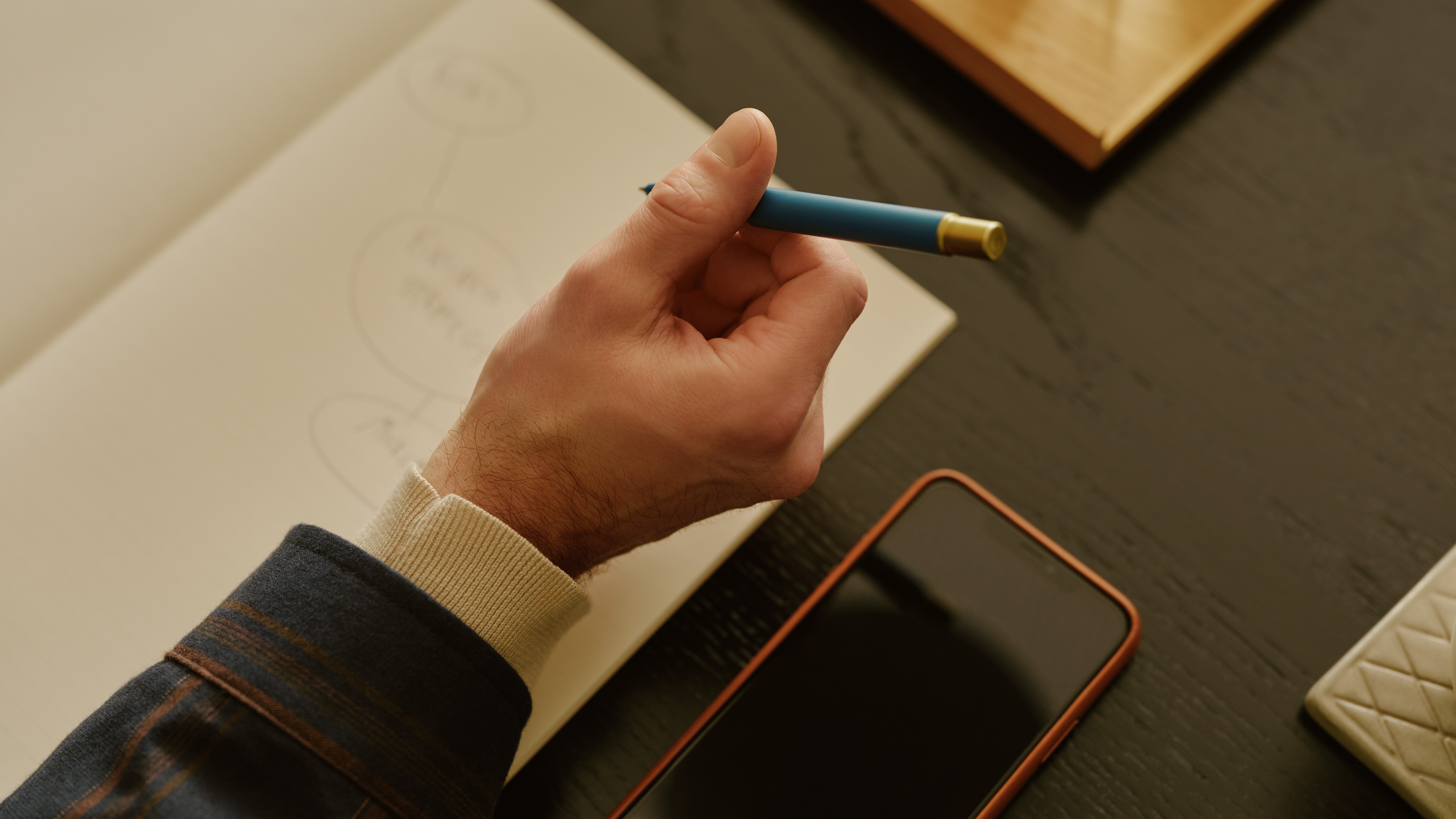 Hand hält blauen Stift über Notizbuch auf dunklem Tisch, daneben Smartphone und Glas Wasser.