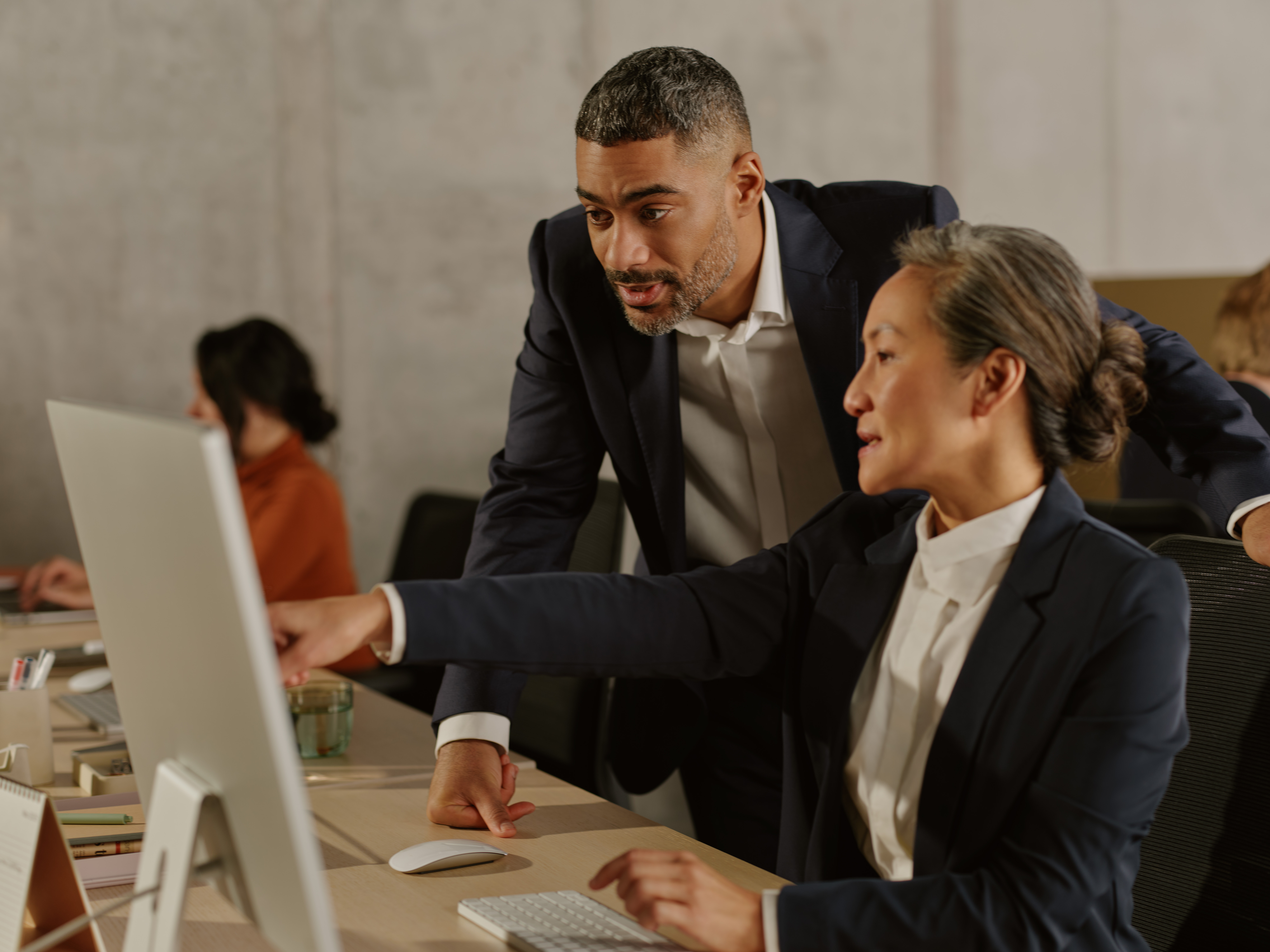 Two colleagues in navy suits collaborate at a desktop computer in a modern office, with coworkers visible in the background.