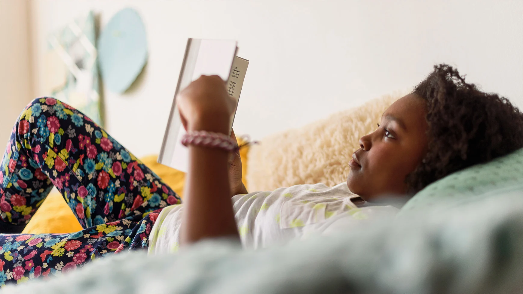 Una niña está recostada leyendo un libro. Viste pantalones floreados y una camiseta blanca.