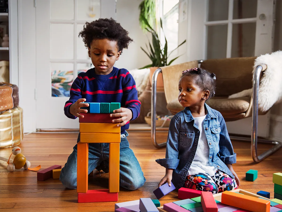 Two siblings are playing with wooden blocks. The older sibling builds a tower while the younger sibling watches. 