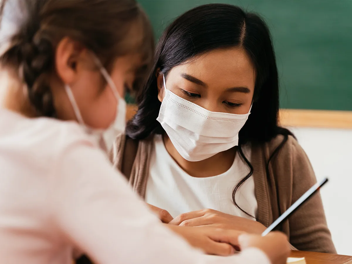 A teacher provides support to a student who is doing schoolwork with a pencil. Both wear facemasks in the classroom.