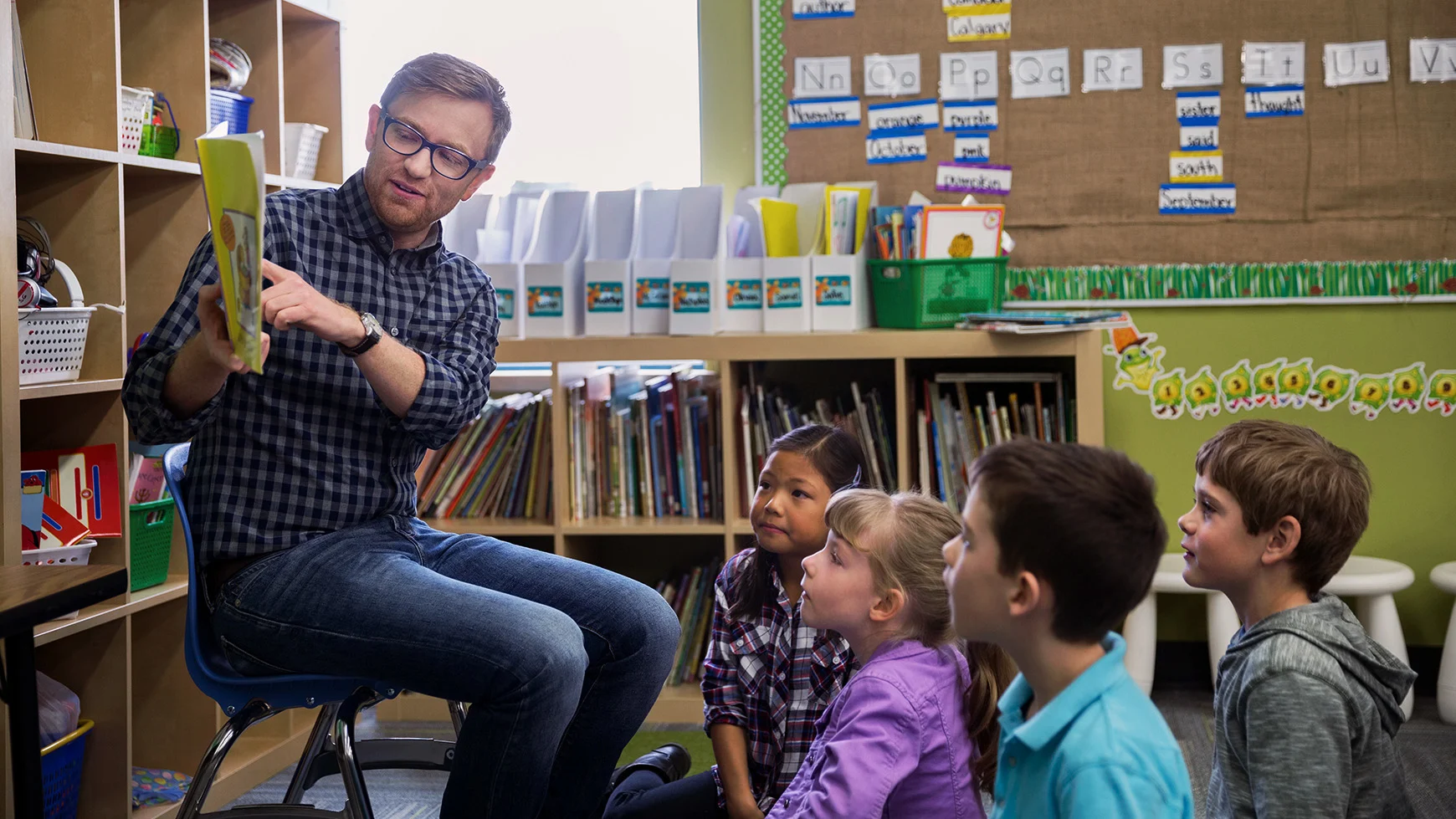 A teacher sits on a chair and holds a book, pointing at a word. The four kids sitting on the floor in the classroom watch. 