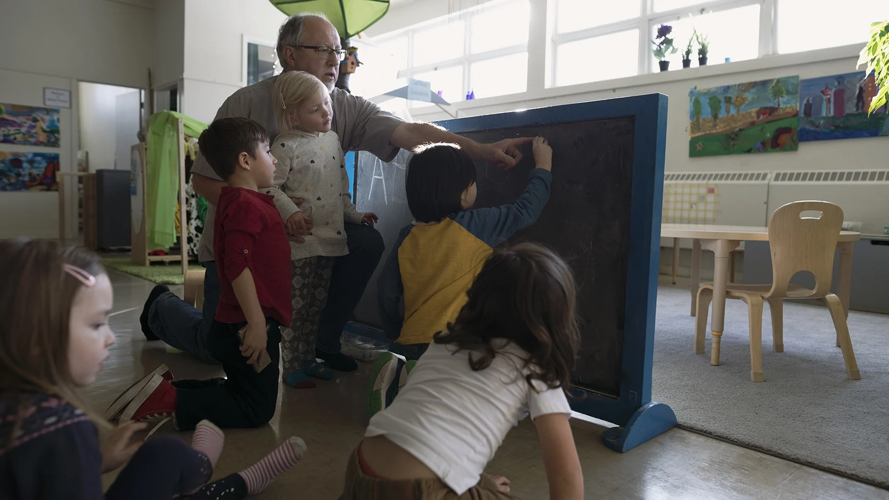 A teacher assists a student in writing on a blackboard. Four other students are watching, while one student is looking away.