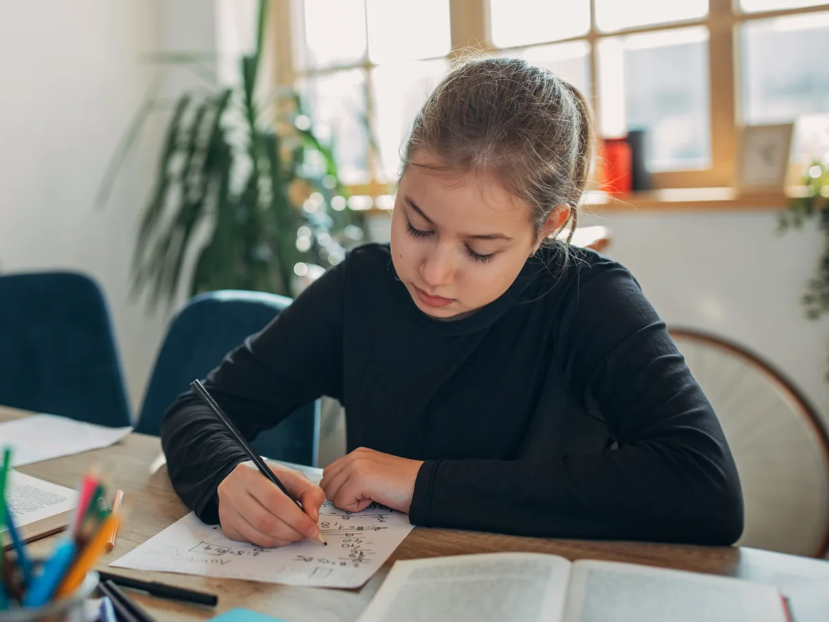 Una estudiante utiliza un lápiz negro para hacer su tarea de matemáticas en una hoja de papel. Sobre el escritorio hay un libro abierto.
