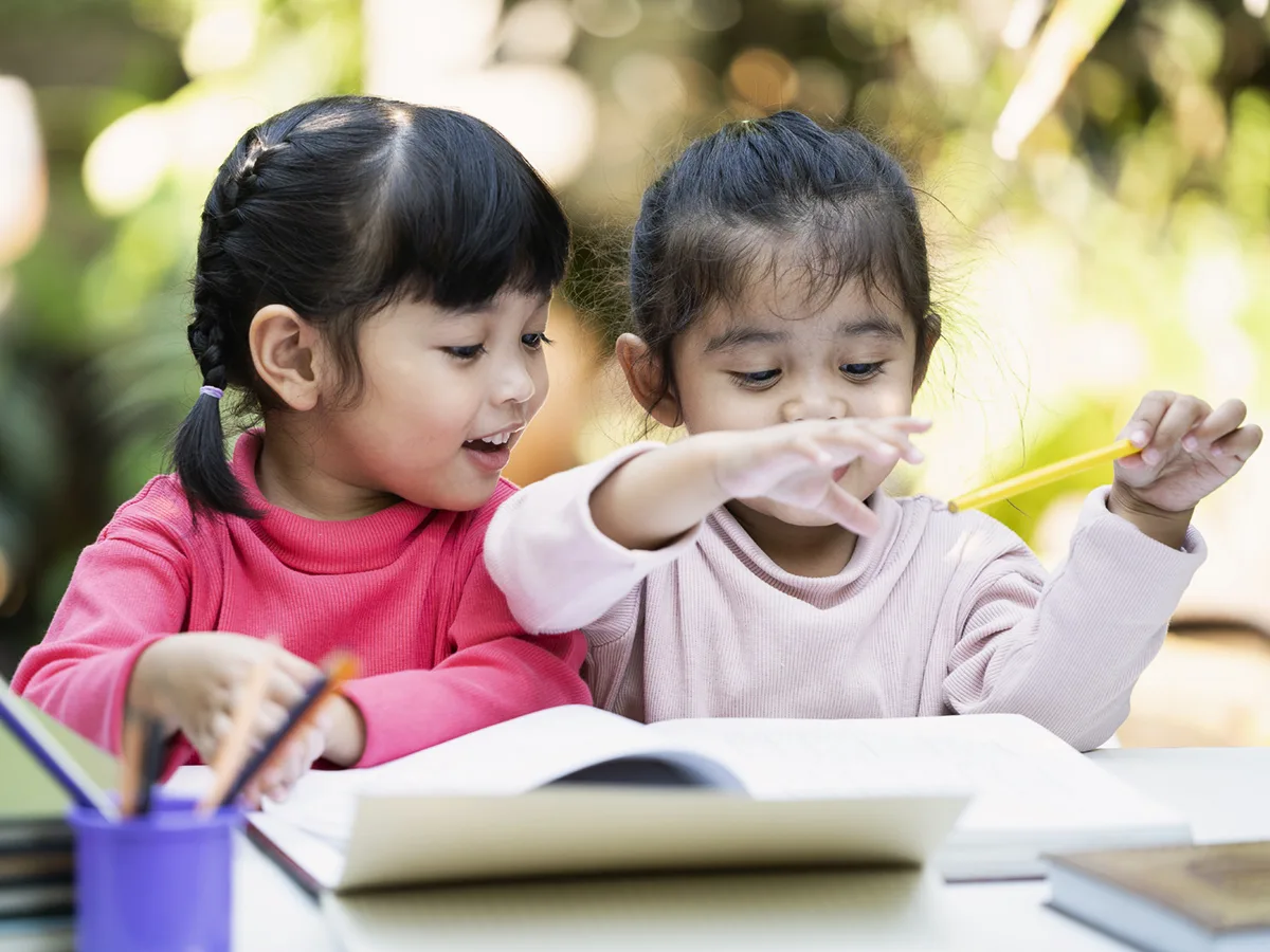 Two young children at a desk — one poised with a pencil above a notebook, the other smiling and observing. 