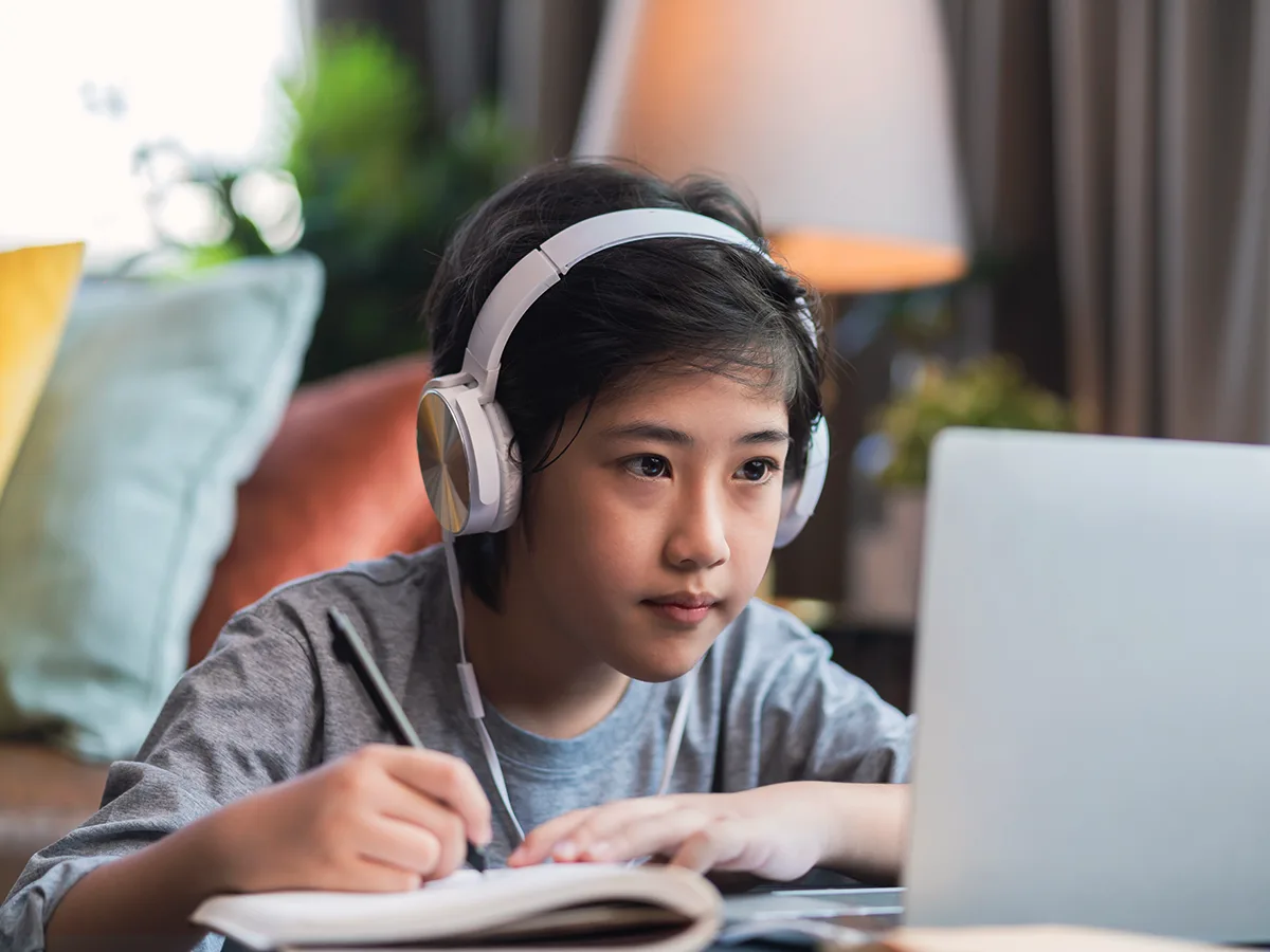 A boy writes in a notebook while looking at a computer with headphones on.