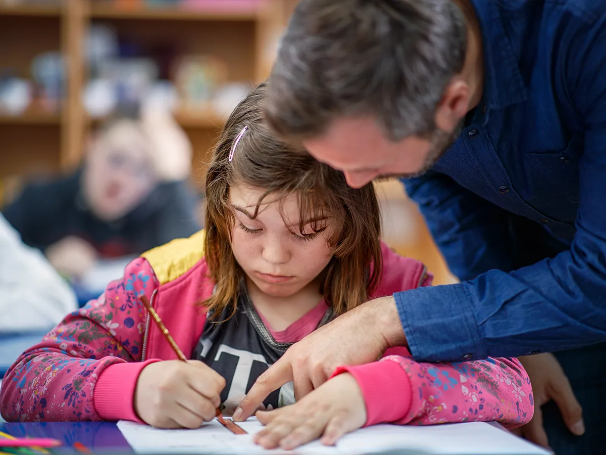 A teacher helps a child who is writing on a piece of paper. 