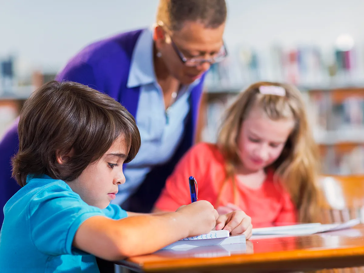 Dos niños están escribiendo sobre papel mientras una maestra observa.